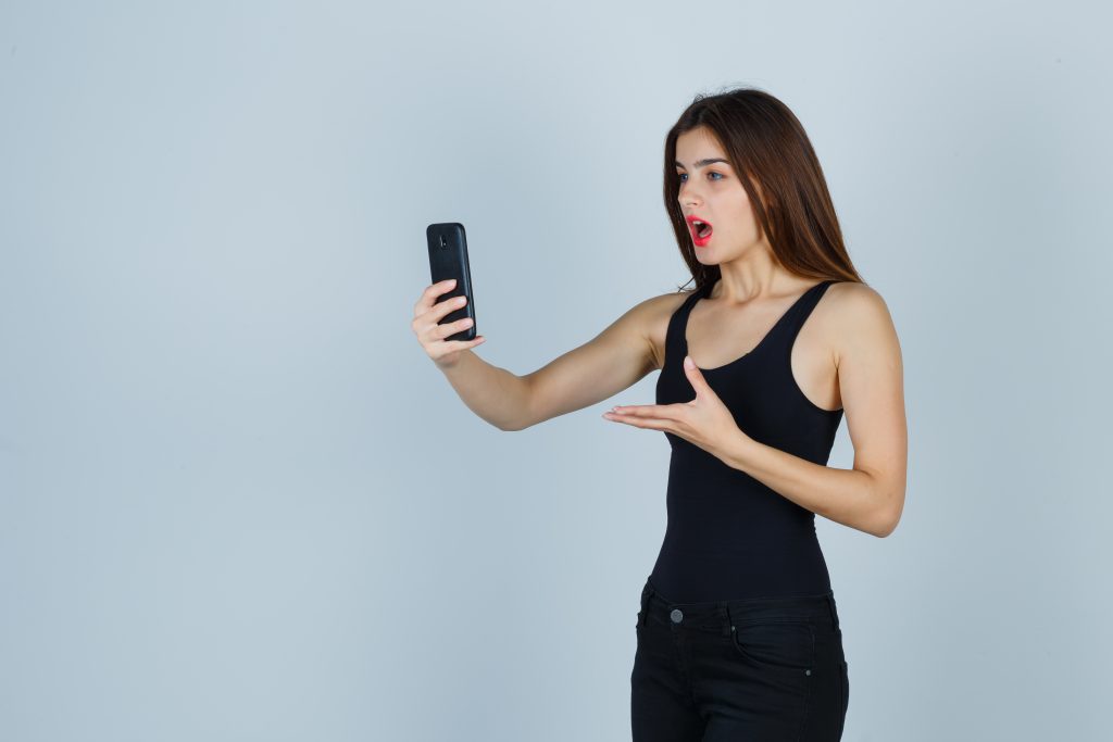 young girl talking to someone via phone, stretching hand toward phone in black top, pants and looking focused. front view.